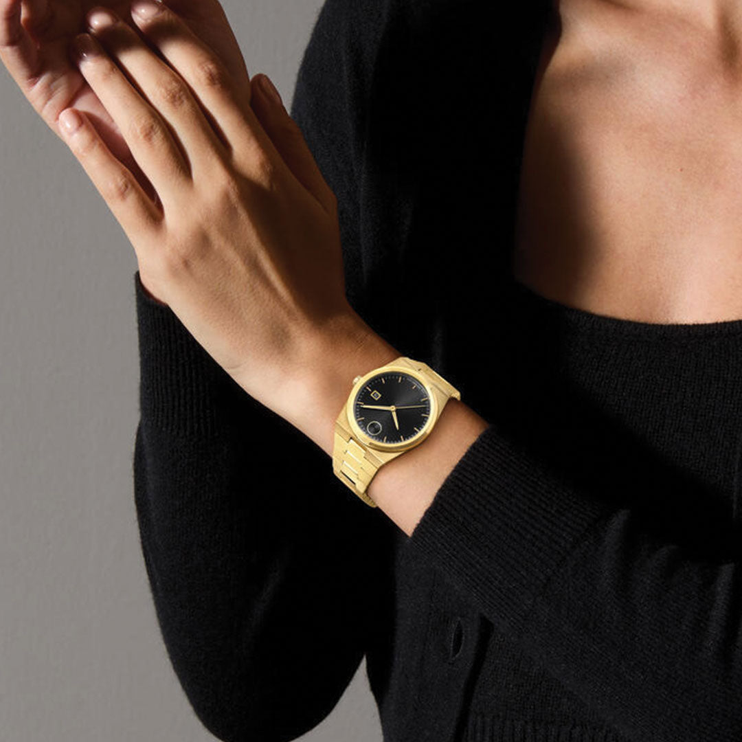 A person in black reveals their left wrist, styled with a Movado BOLD Quest Quartz 40mm Watch featuring an integrated bracelet and bold black sunray dial. Their hands gently touch against a neutral background.