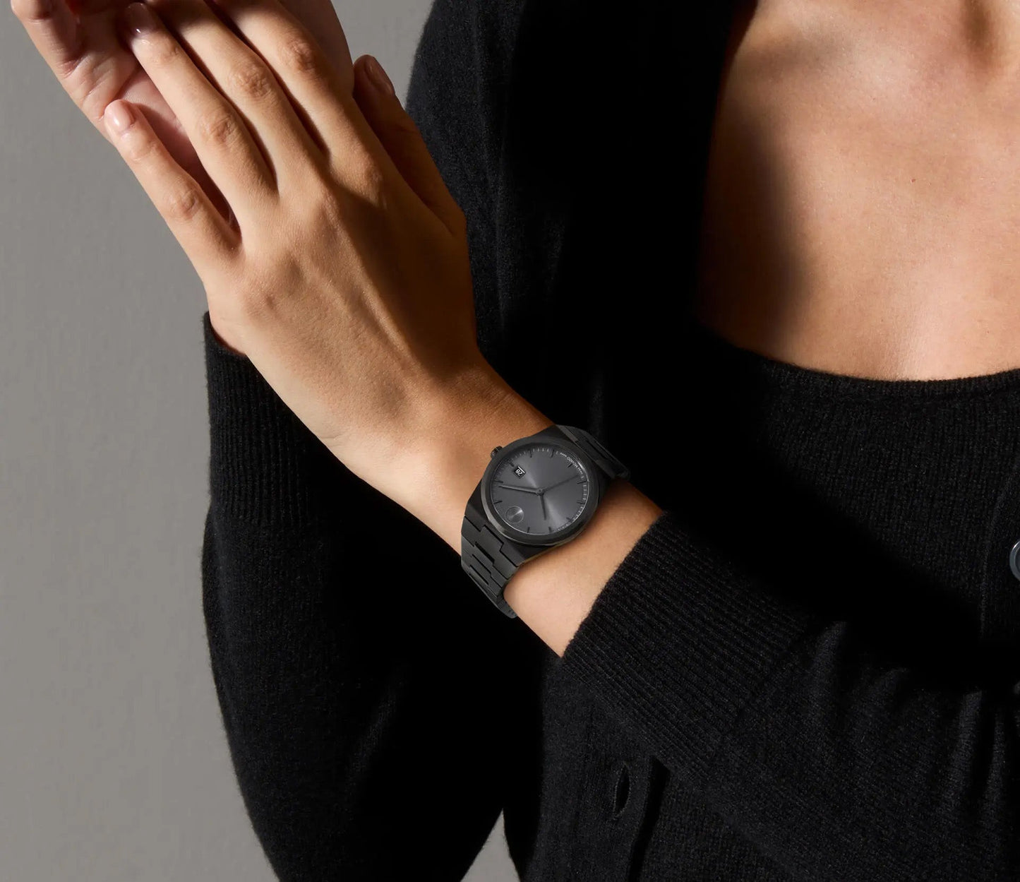 A person wearing a Movado BOLD Quest Quartz 40mm Watch, featuring an integrated bracelet and minimalist design, adjusts their clasped hands. They are dressed in a black cardigan and surrounded by a neutral background, evoking the precision and style of Movado.