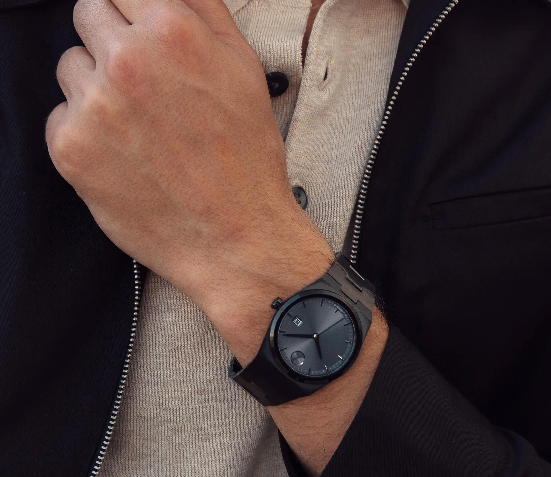 A person wearing the Movado BOLD Quest Quartz 40mm Watch on their left wrist is dressed in a dark jacket and beige shirt, with the wrist resting on their chest. The watch, featuring an integrated bracelet and a minimalistic design from Movado, boasts a clean black look with a dark dial that enhances its seamless style.