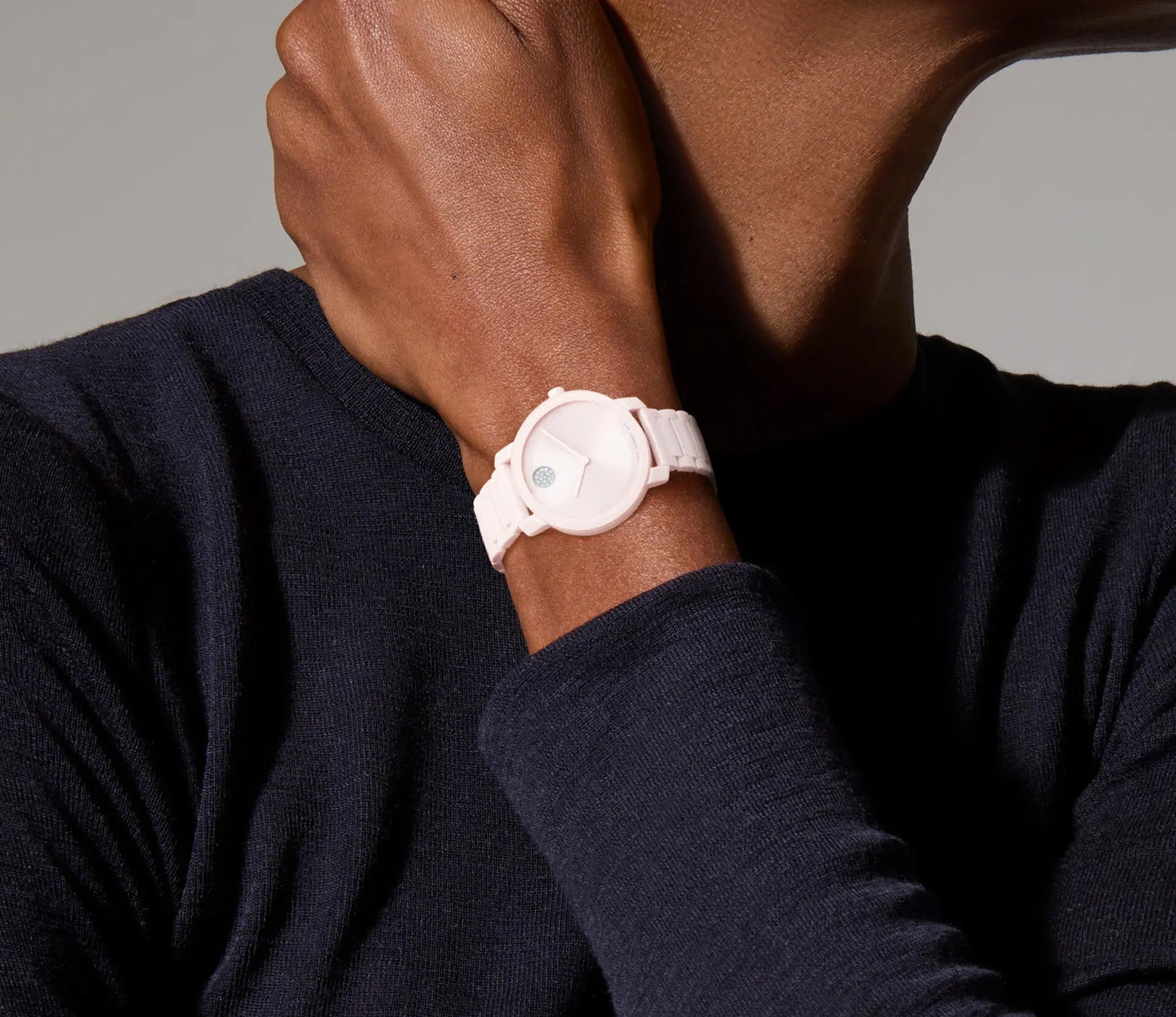 Dressed in a black long-sleeve shirt, an individual showcases a white Movado BOLD Evolution 2.0 Quartz 34mm Watch with an H-link bracelet. The watch has a minimalist face featuring a micro-textured dial and a small blue circle accent. By holding their wrist near their face, they gracefully emphasize the elegance of the watch's design from Movado.