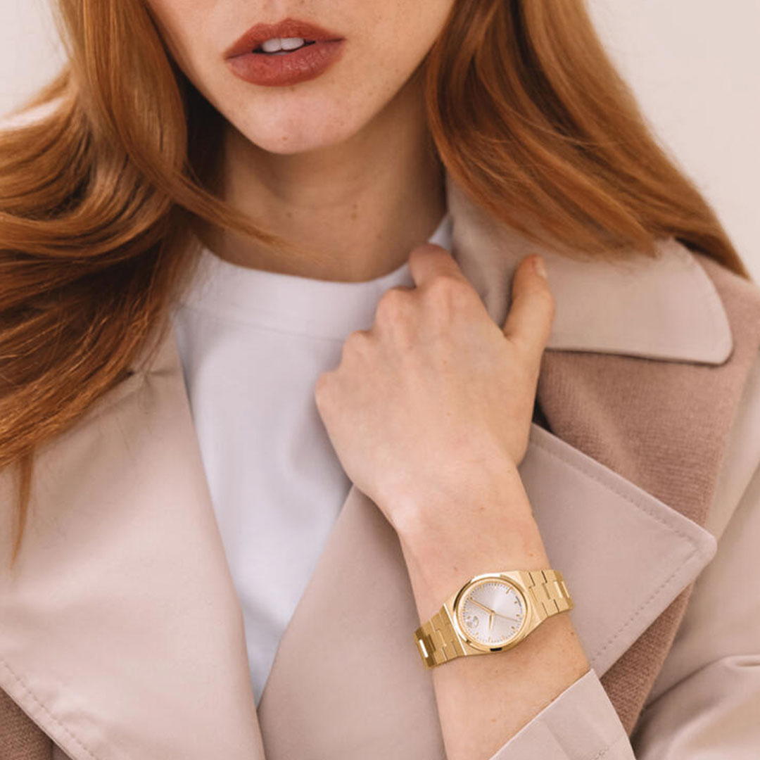 A woman with long red hair wears a light beige coat over a white top, accessorized with a Movado BOLD Quest Mini Quartz 28mm watch. She touches her coat collar, showing off the elegant Movado timepiece on her wrist as her face remains partially visible.