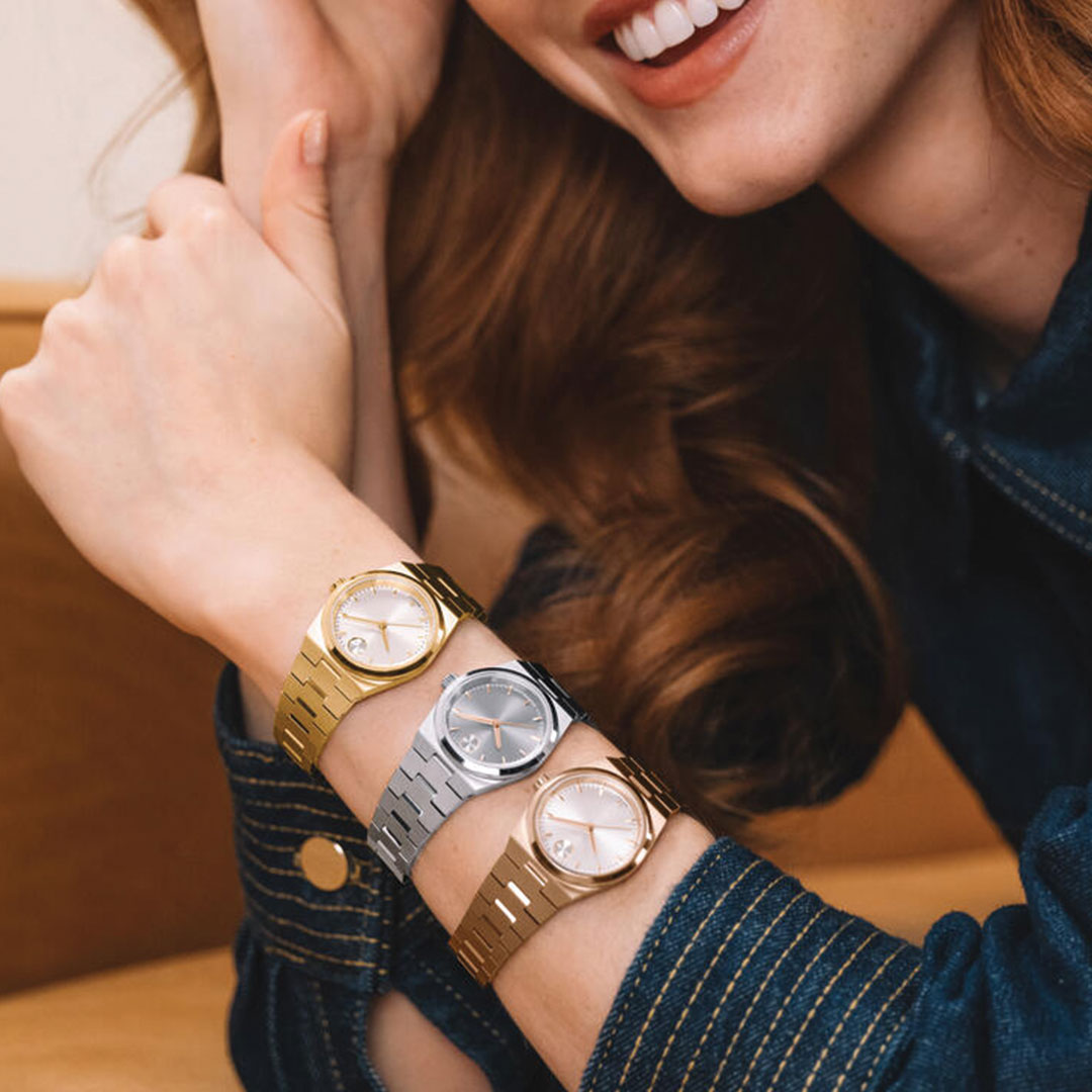 A smiling person in a dark denim jacket shows off three wristwatches—including a 28mm Movado BOLD Quest Mini Quartz with a champagne dial—stacked on their arm in gold, silver, and two-tone styles.