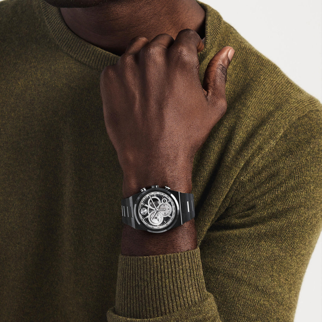 A person wearing an olive green sweater rests their chin on their hand, showcasing the Movado BOLD Quest Chronograph Quartz 40mm Watch by Movado, featuring a detailed silver skeleton dial and a sleek stainless steel bracelet.