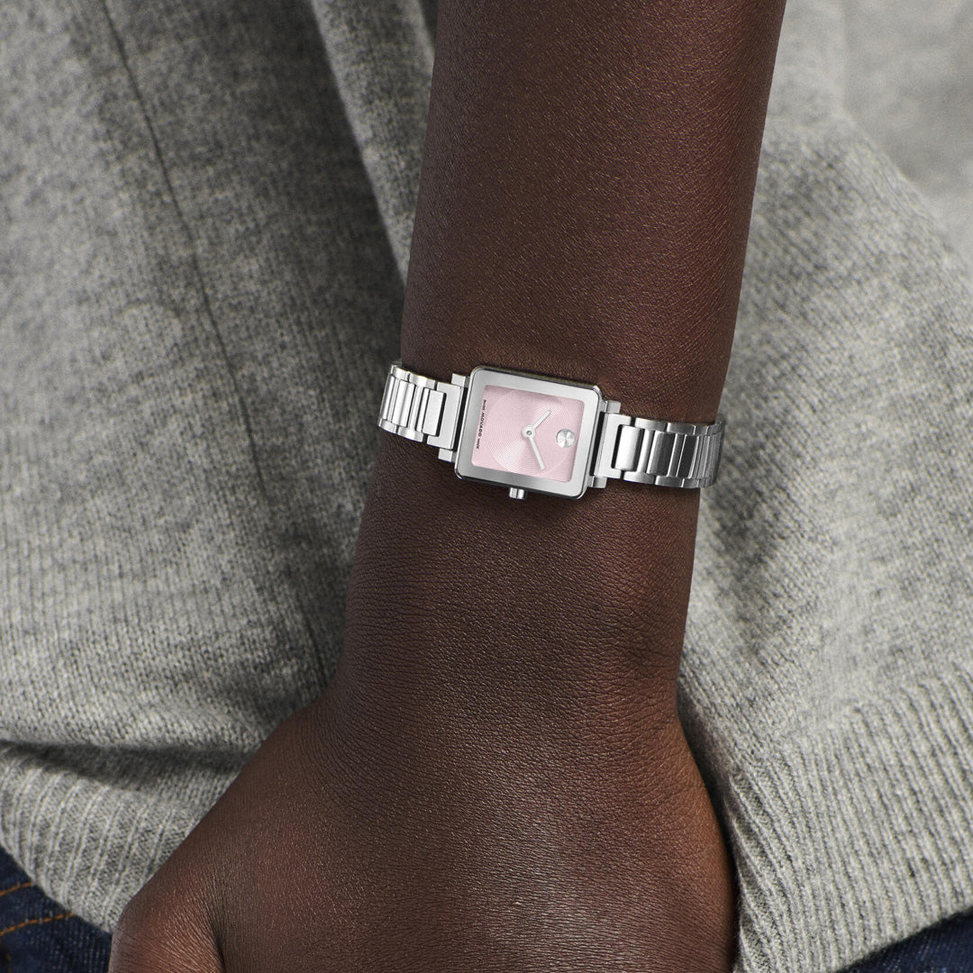 A person wearing the Movado BOLD Evolution 2.0 Mini Quartz 19mm x 22.5mm Watch, featuring a silver metal band and a square pink microtextured dial, shown against a gray sweater.