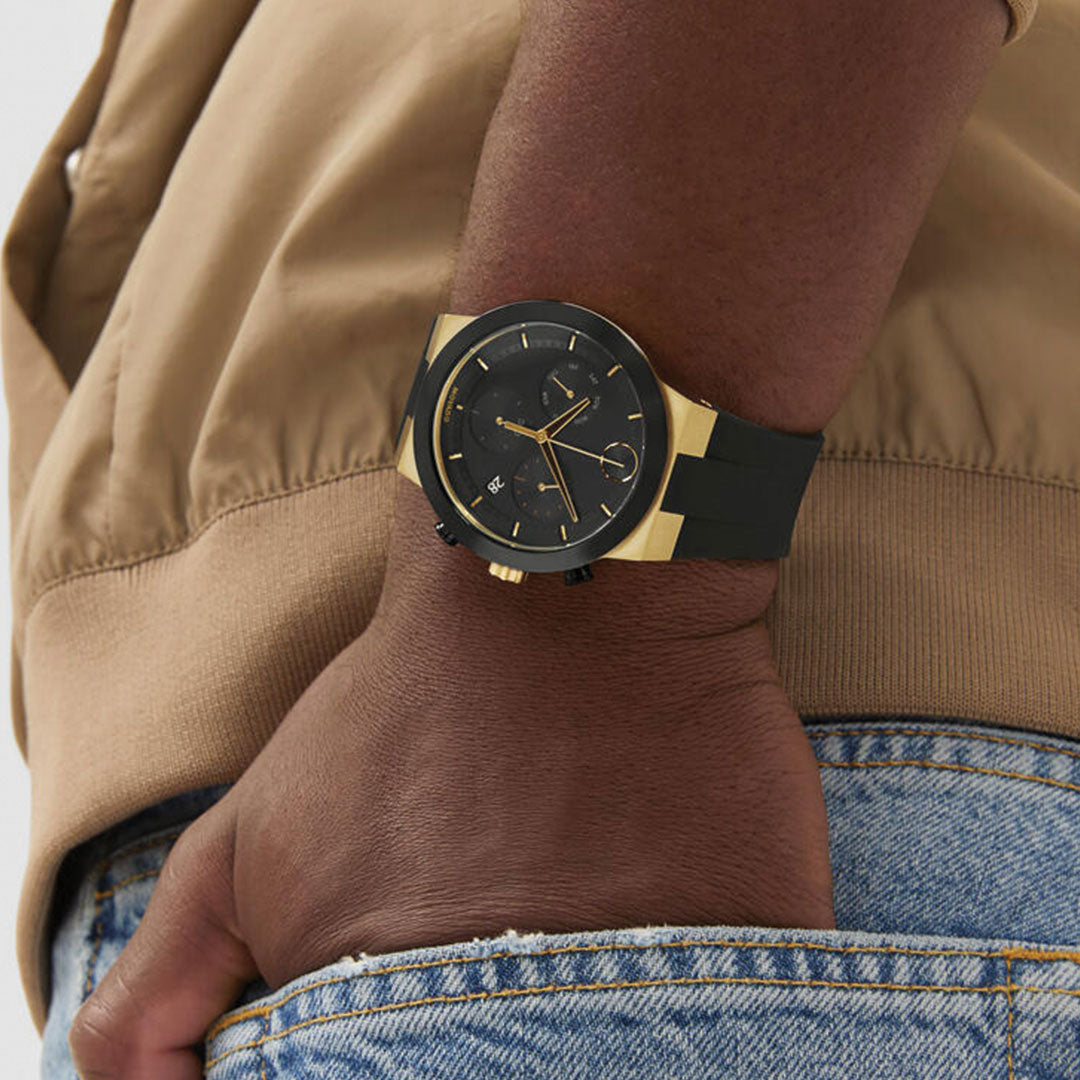 A person in a tan jacket and blue jeans wears a Movado BOLD Fusion Chronograph Quartz 44.5mm Watch with a round face and black strap on their left wrist, hand relaxed and resting near their hip.
