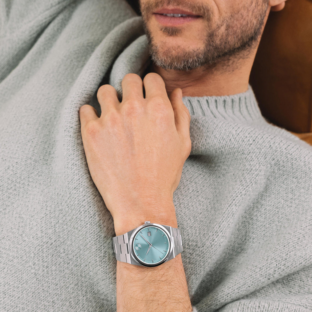 A man wearing a light gray sweater is reclining, displaying a Movado BOLD Quest Quartz 40mm Watch with a pale blue face and stainless steel case on his left wrist. Only his lower face and hand are visible.