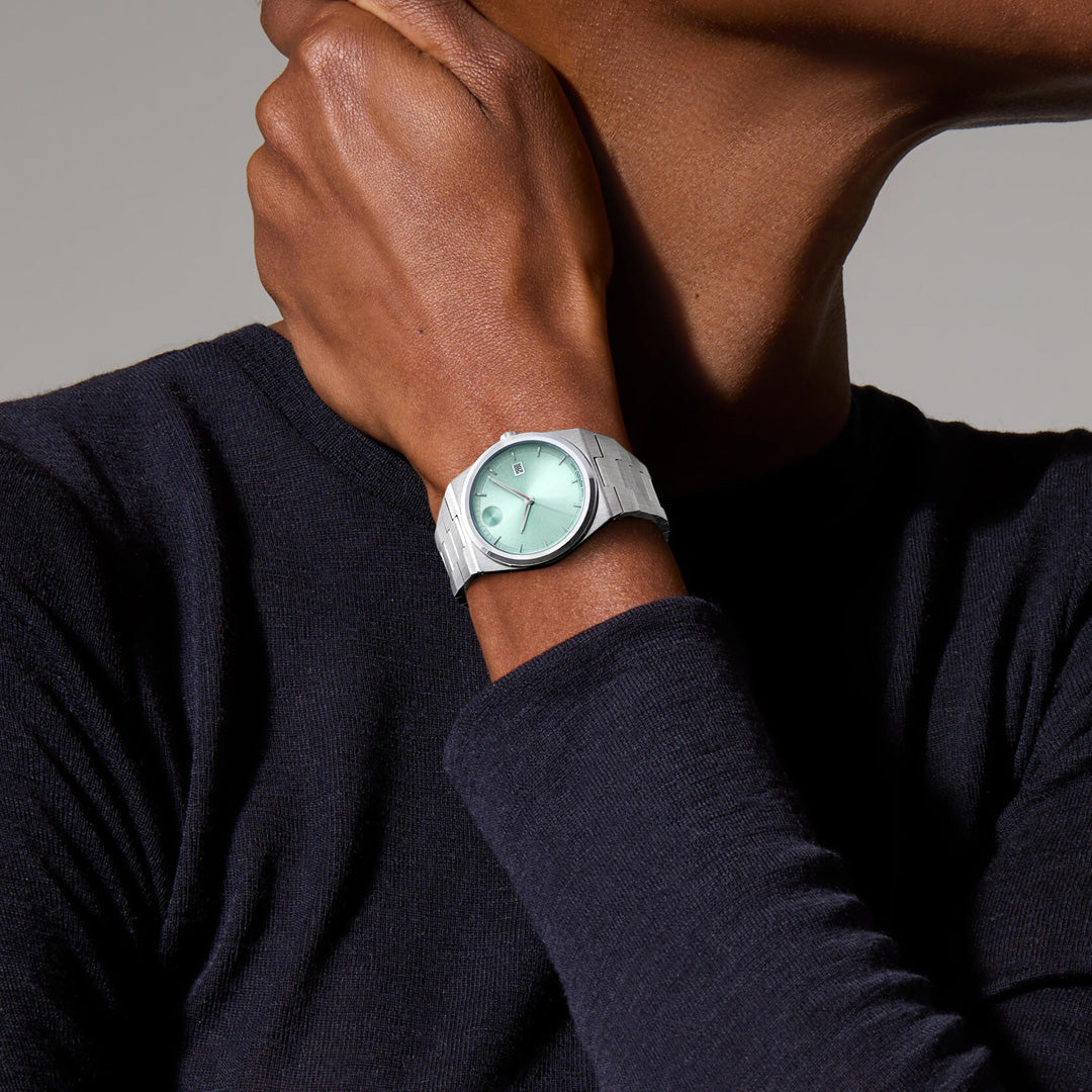 A person in a dark long-sleeve shirt holds their neck, showing off the Movado BOLD Quest Quartz 40mm Watch by Movado, featuring a mint green face and stainless steel construction. The neutral background is softly blurred.