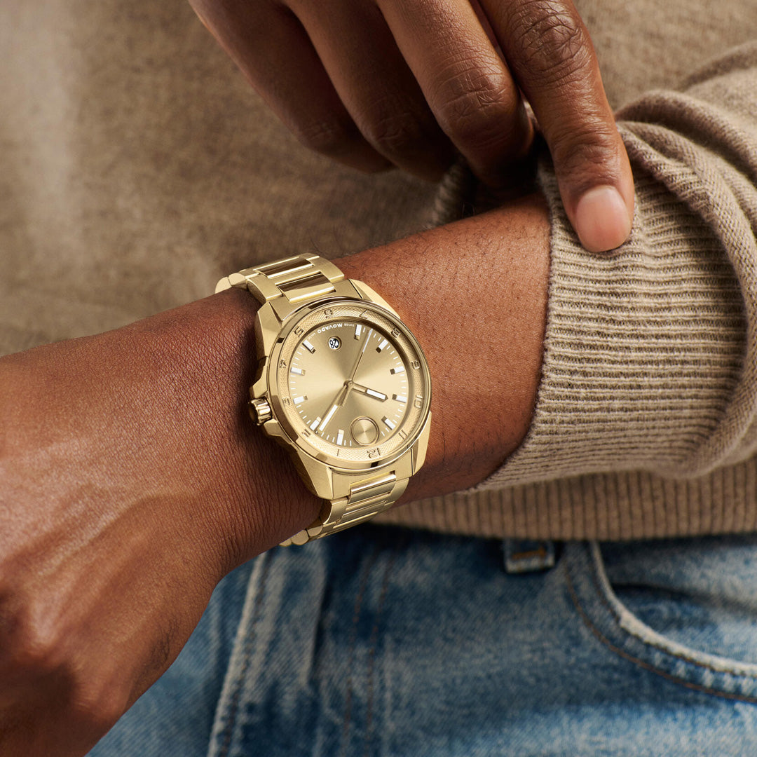 A person wearing a beige sweater and blue jeans points at their wrist, which features the Movado BOLD Verso S Quartz 42mm Watch in yellow gold with a Swiss-made movement.