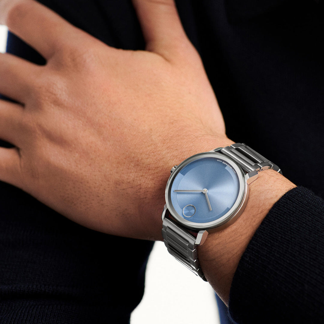 A person wearing a Movado BOLD Evolution 2.0 Quartz 40mm Watch, featuring a silver case and a blue, micro-textured dial without numbers, rests their hand on their opposite forearm.