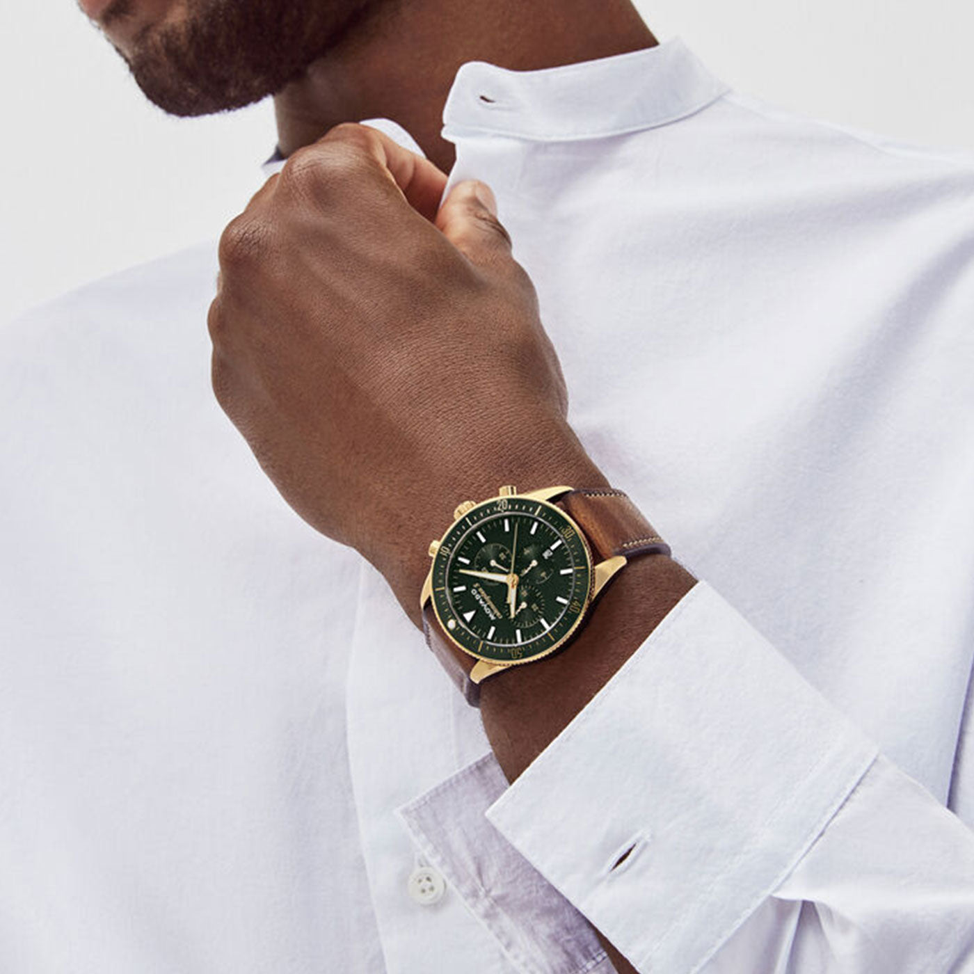 A man in a white shirt adjusts his collar with one hand. His wrist features a Movado Heritage Series Calendoplan S Quartz 42mm Watch, showcasing a gold case and brown leather strap, complemented by a green face with three sub-dials. The setting is minimalistic and elegant.