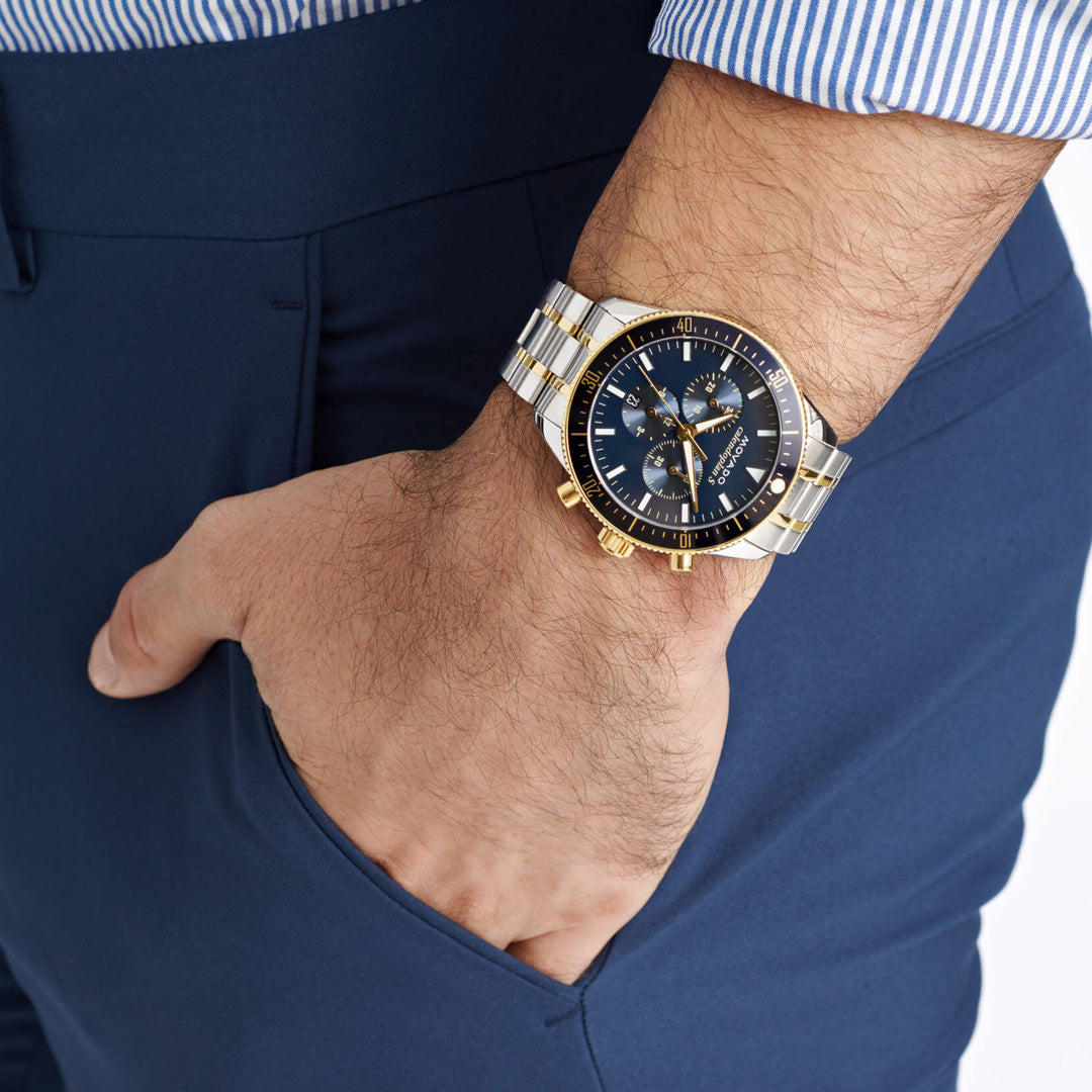 A person wearing the Movado Heritage Calendoplan S Chronograph Quartz 42mm Watch by Movado, featuring a blue and gold ion-plated case and a blue dial, is dressed in a blue and white striped shirt with navy pants, their hand casually resting in their pocket.