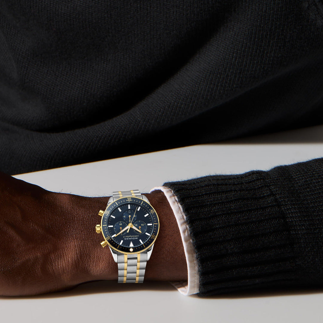 A person wearing a black sweater and a Movado Heritage Calendoplan S Chronograph Quartz 42mm Watch with a silver and gold ion-plated band and black face rests their arm on a white surface.