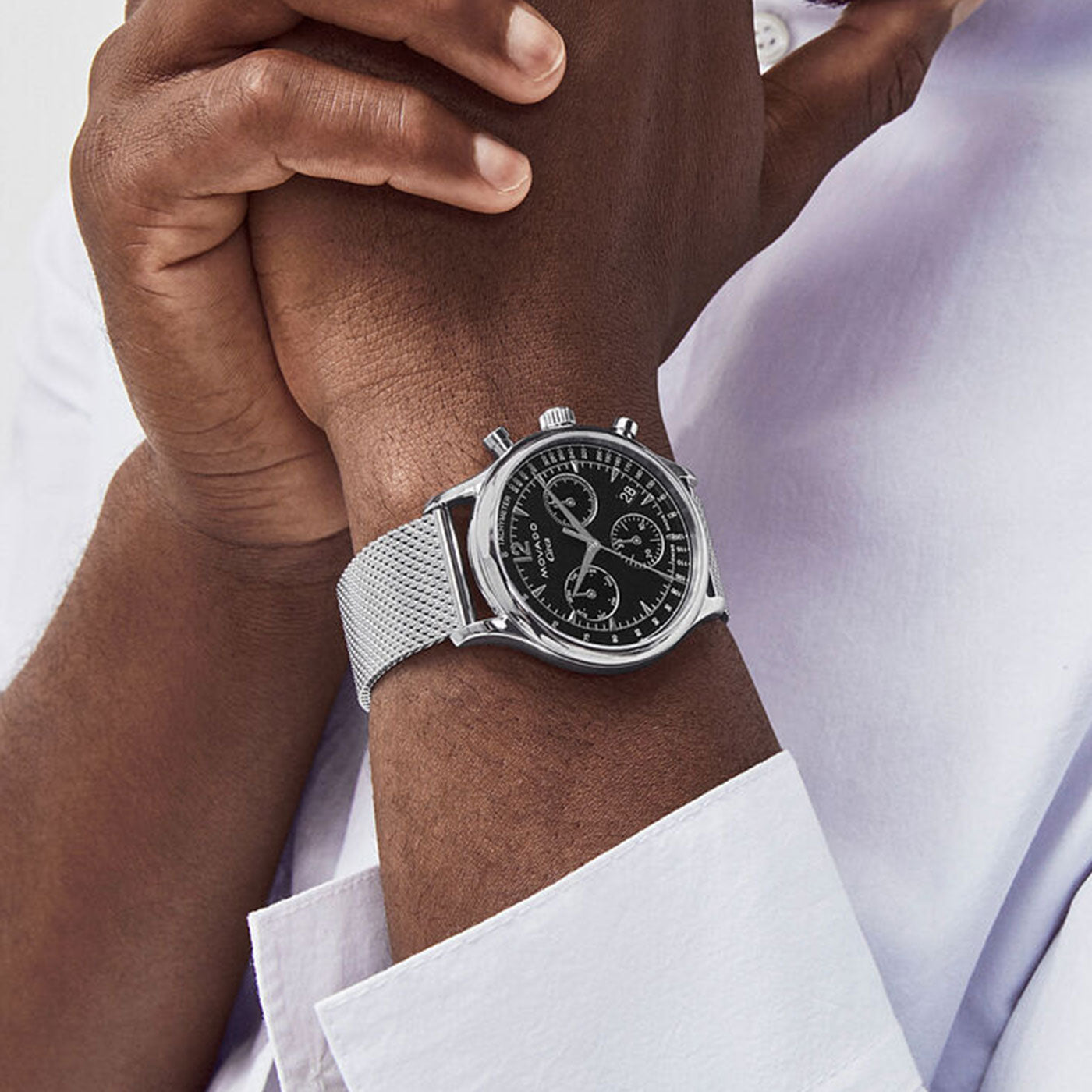 A person is wearing the Movado Heritage Series Circa Quartz 43mm Watch, featuring a sleek stainless steel design with a striking black dial and mesh band. Dressed in a white shirt, their hands are clasped together, prominently displaying the eye-catching timepiece from Movado.