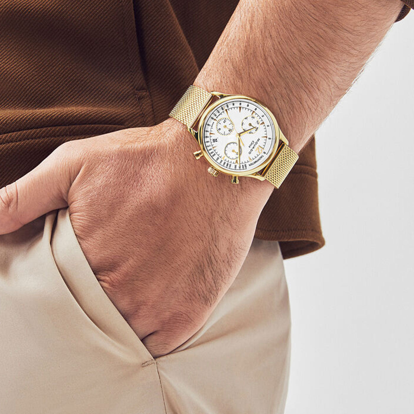 A person is wearing a Movado Heritage Series Circa Quartz 43mm watch from the brand Movado, featuring a gold mesh strap and a white dial with three subdials that evoke a mid-century design. Their hand is tucked into the pocket of beige pants, and they are donning a brown shirt. The watch exudes a sleek, modern aesthetic.