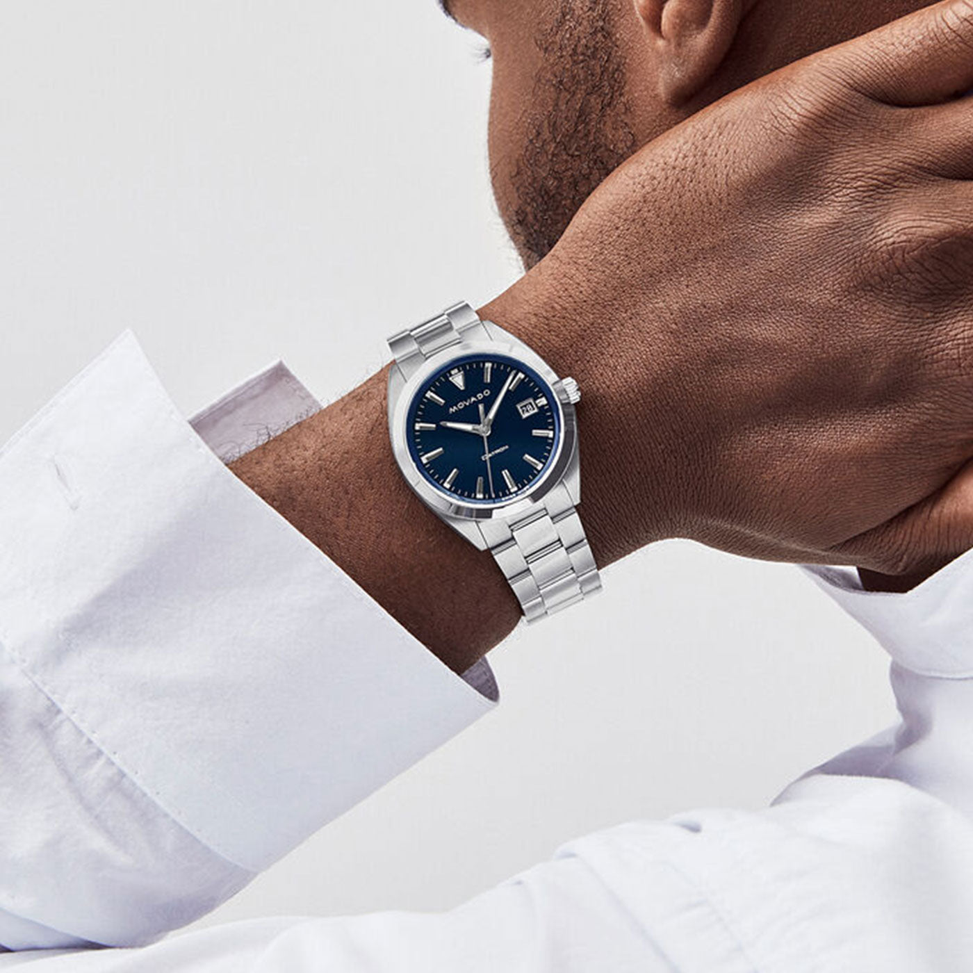 A person is wearing a Movado Heritage Series Datron Quartz 39mm watch with a blue face, paired with a partially visible white shirt sleeve. This retro-cool design from the renowned Movado brand features a classic look from the Datron Collection, complete with a stainless steel bracelet that elegantly showcases time and date.