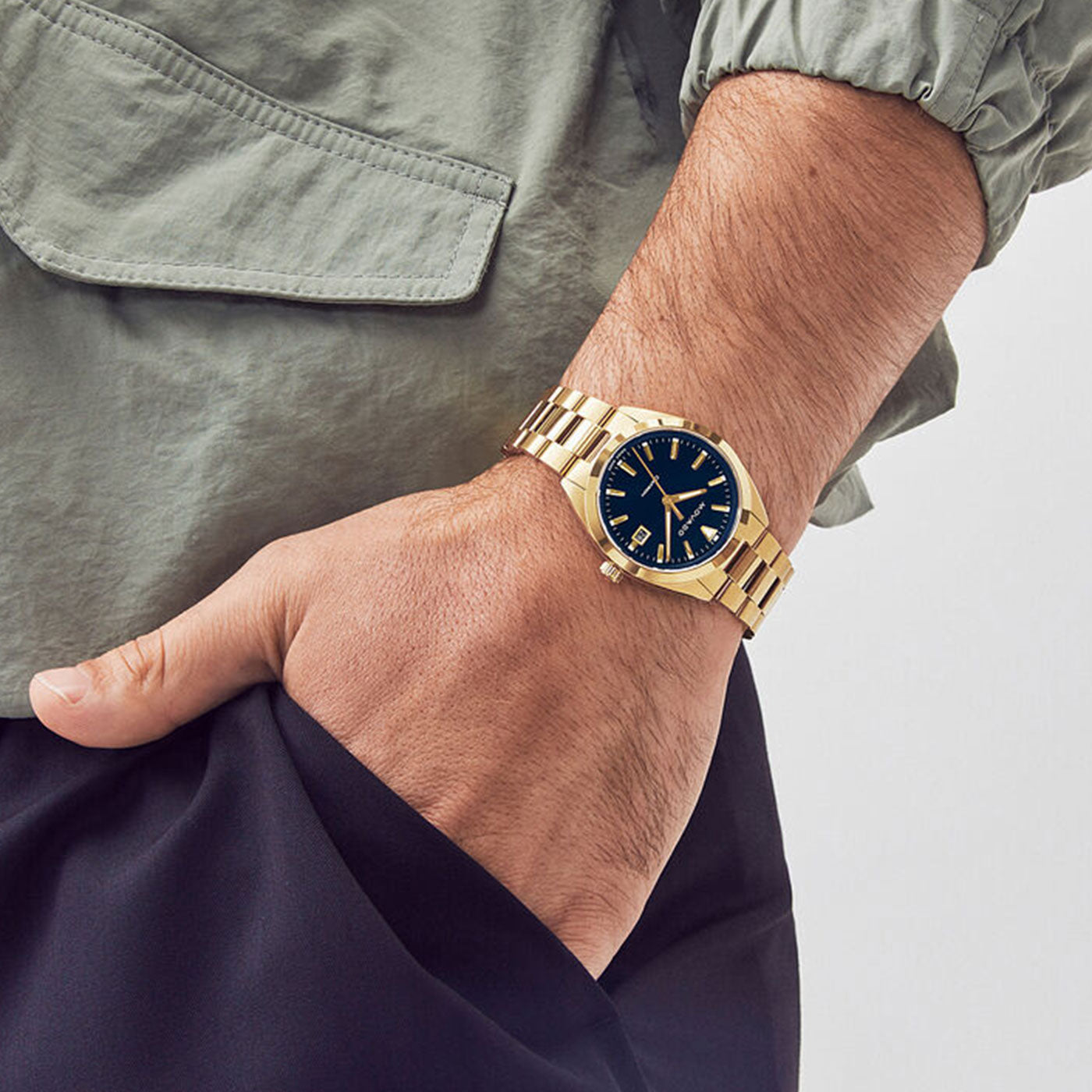 A person wears a Movado Heritage Series Datron Quartz 39mm watch with a dark blue face. This yellow gold wristwatch from the Movado brand features Swiss Super-LumiNova-enhanced gold hour markers and a metal strap. Their hand is casually tucked into the pocket of dark pants, paired with a stylish light green jacket.