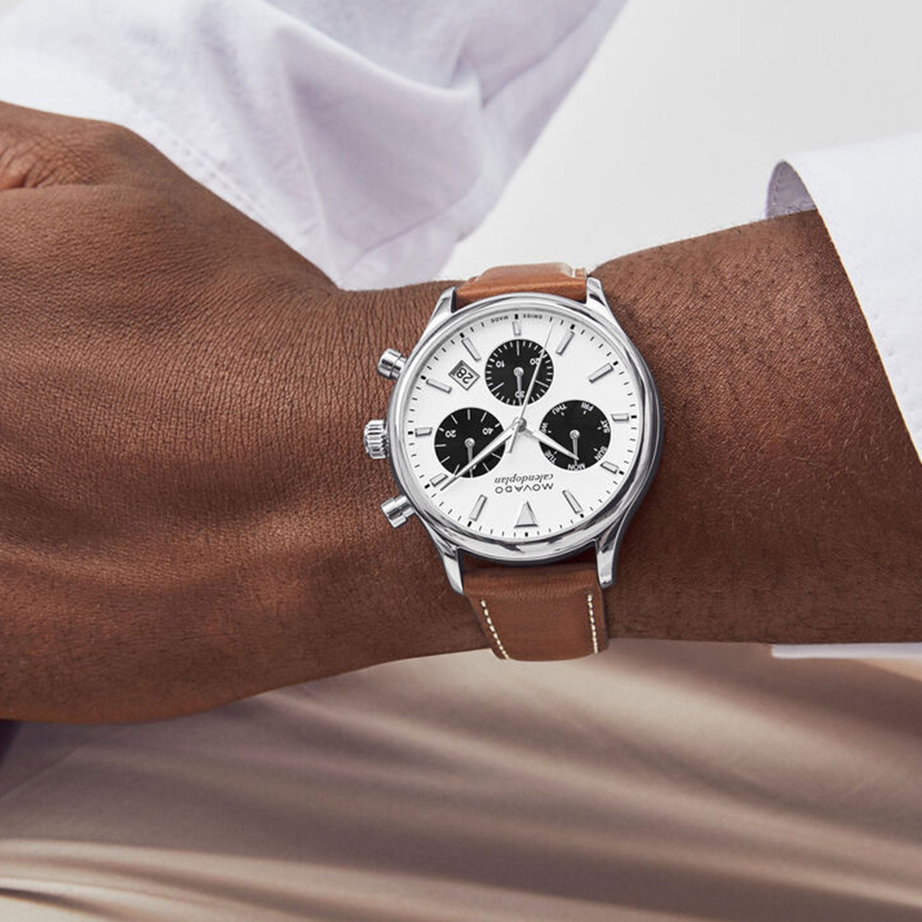 Close-up of a person wearing a white dress shirt with a silver and brown wristwatch on their left wrist. This Movado Heritage Series Calendoplan Quartz 43mm watch features a white face with dark subdials, encased in polished stainless steel, and paired with a brown leather strap, resting on their lap.