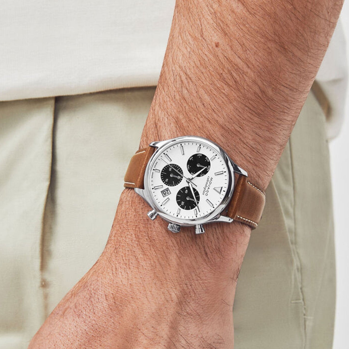 A close-up shot captures a person wearing the Movado Heritage Series Calendoplan Quartz 43mm Watch, which boasts a brown leather strap and a sleek stainless steel case. The timepiece features a white face with three black subdials and a date display. The individual is styled in light-colored pants and a crisp white shirt, perfectly complementing their sophisticated chronograph watch.