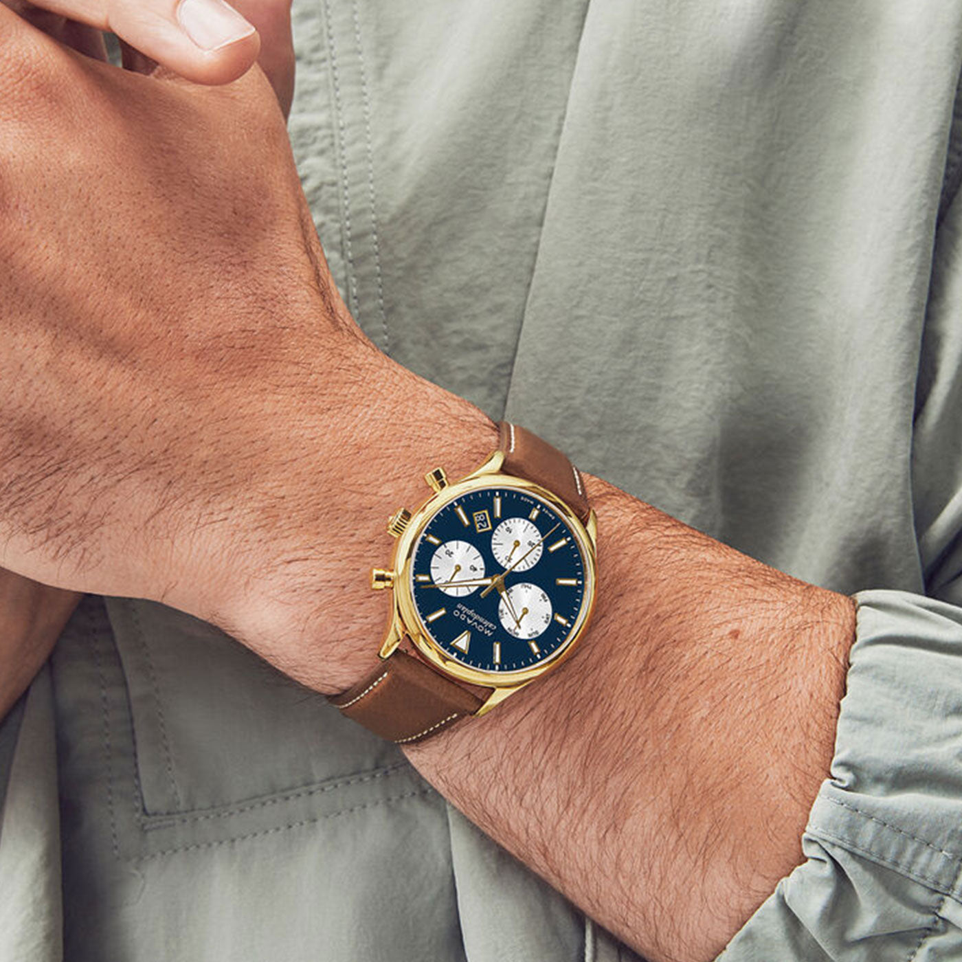 A person is sporting a Movado Heritage Series Calendplan Quartz 43mm watch, characterized by its gold casing and brown leather strap. The timepiece showcases a dark blue dial with three subdials and gold buttons on the side. Complementing the watch stylishly, the individual is wearing a light gray long-sleeve shirt.