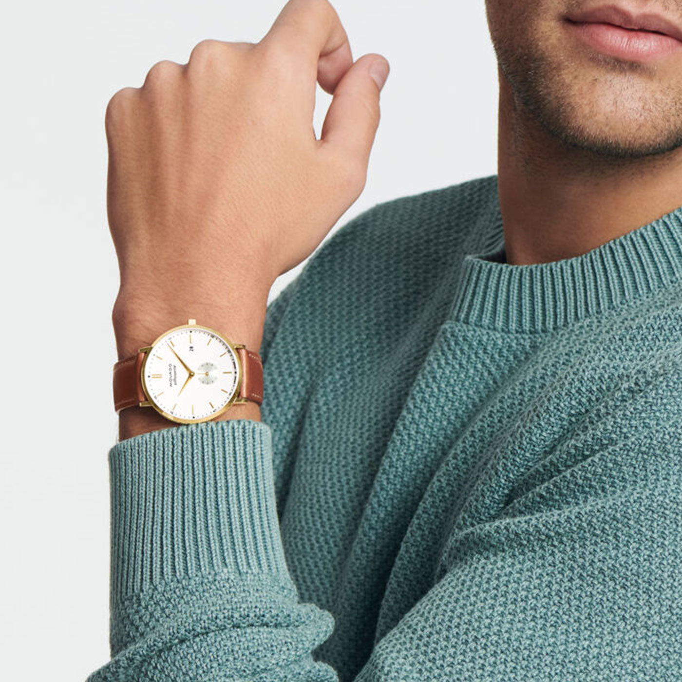 A person in a teal sweater is shown from the shoulder up, with their left hand near their chin, wearing a Movado Heritage Series Silhouette Quartz 40mm Watch featuring a leather strap and yellow gold finish. The background is a plain light gray.