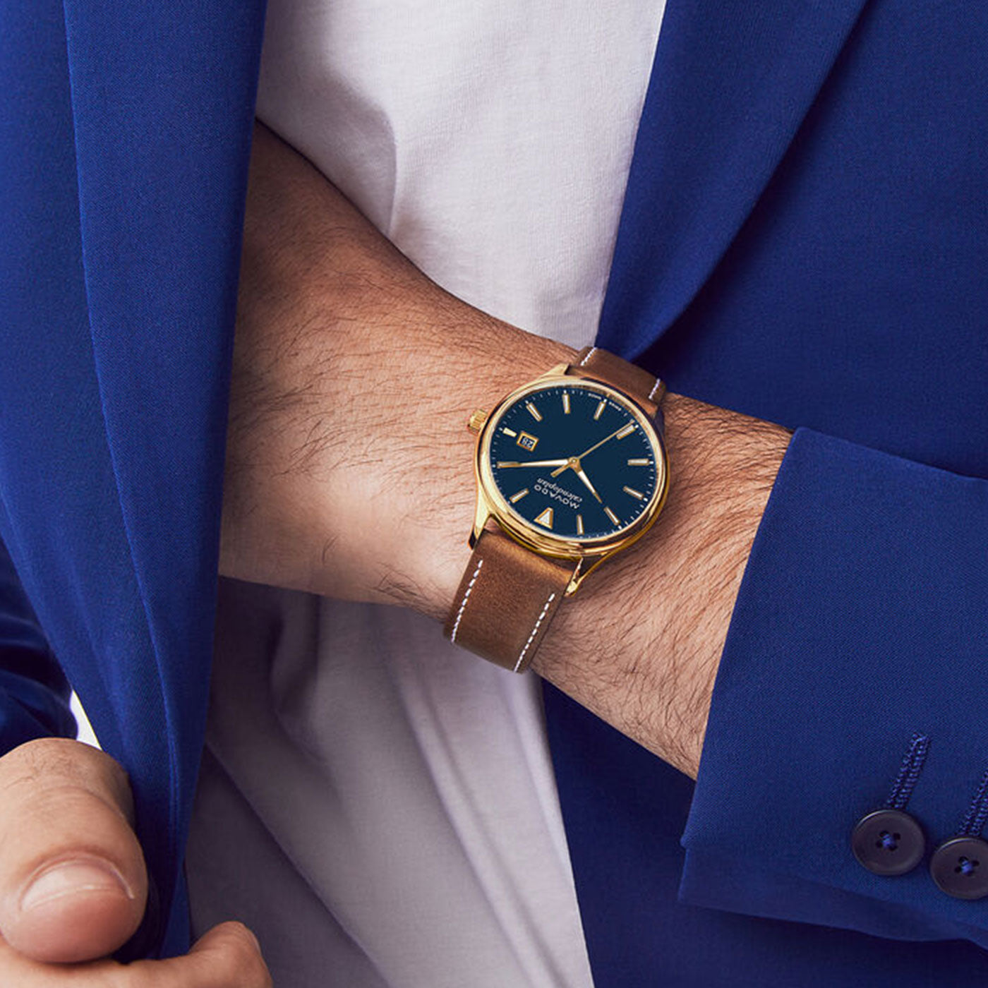 A person wearing a blue suit adjusts their jacket, revealing a Movado Heritage Series Calendoplan Solar Watch with a brown leather strap and gold case. The watch features a black dial with gold markers, echoing a mid-century design that captures the classic elegance reminiscent of the Movado Calendoplan style.