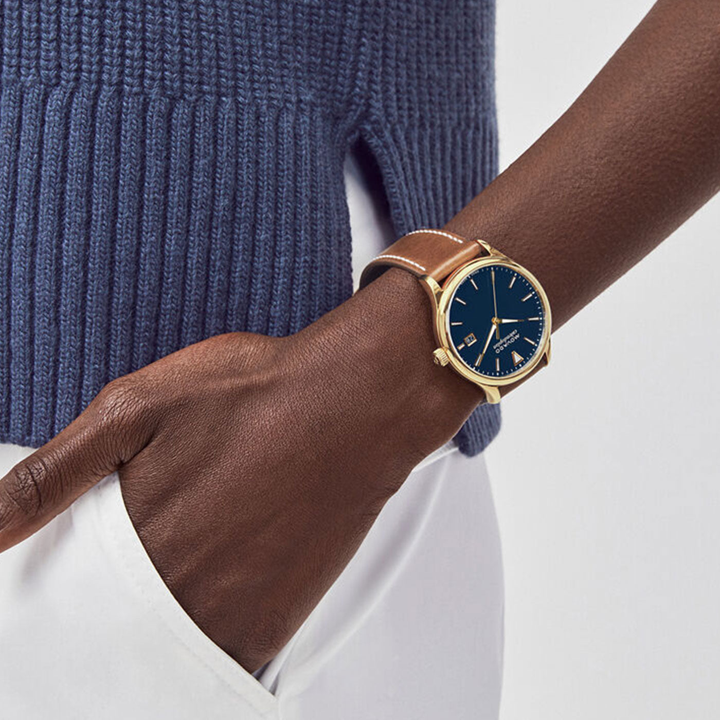 A person wearing a blue sweater and white pants has their left hand in their pocket, showcasing the Movado Heritage Series Calendoplan Solar 40mm Watch. This wristwatch features a yellow gold ion-plated stainless steel case, dark blue face, and brown leather strap, embodying the elegance of mid-century design.