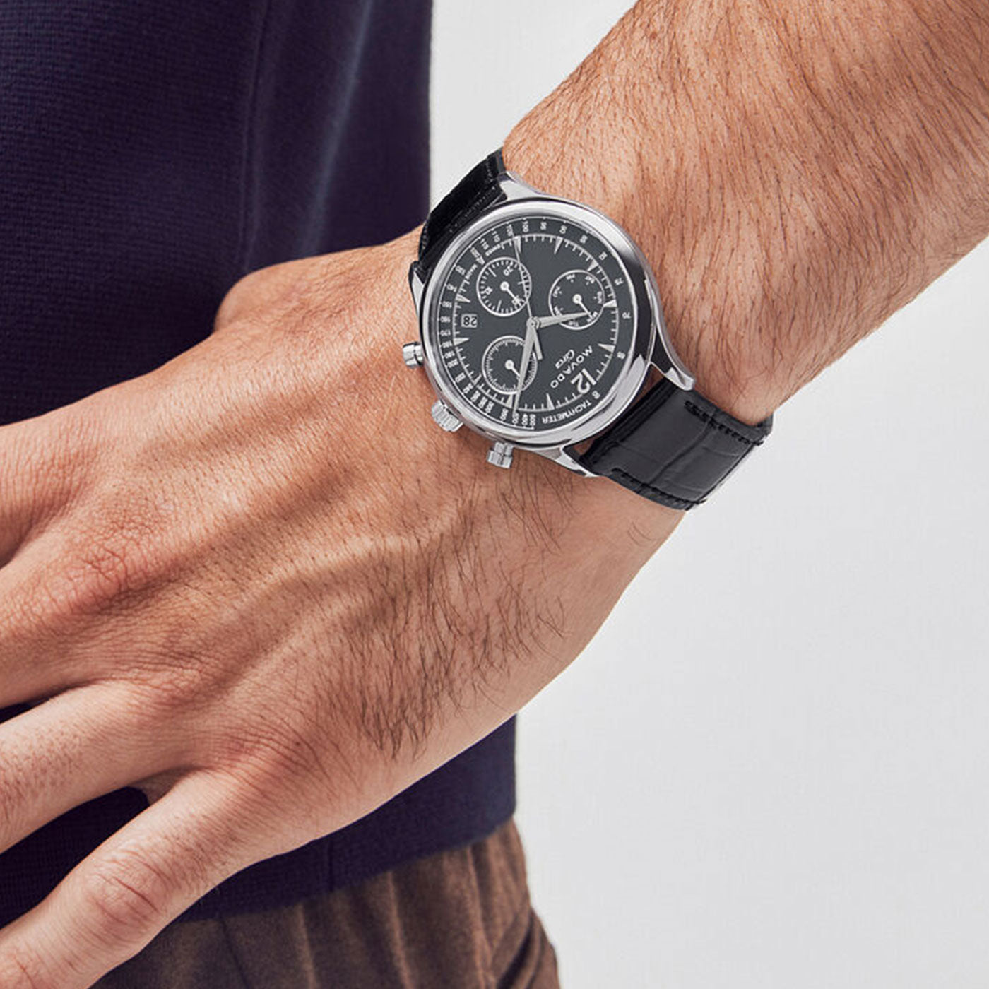 A close-up shot reveals a person's wrist adorned with the Movado Heritage Series Circa Quartz 43mm Watch, featuring a sleek black and silver design with a leather strap. The stainless steel face of this Movado watch showcases multiple dials and numbers. The individual is wearing a dark shirt paired with brown pants.