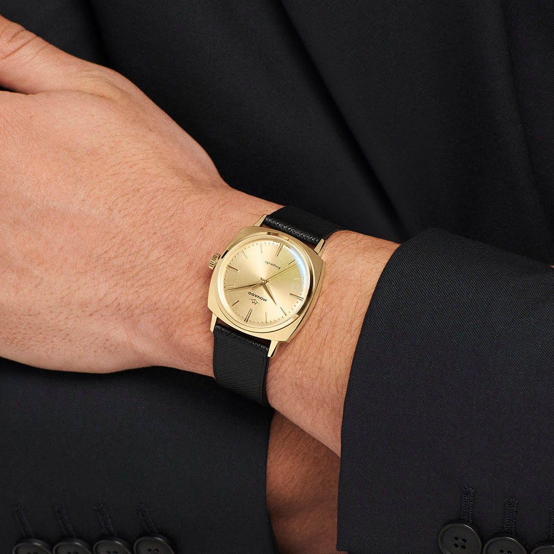 Gold-toned Swiss made Movado Heritage Kingmatic Automatic 35mm Watch with a sunray dial and black strap worn on a wrist.