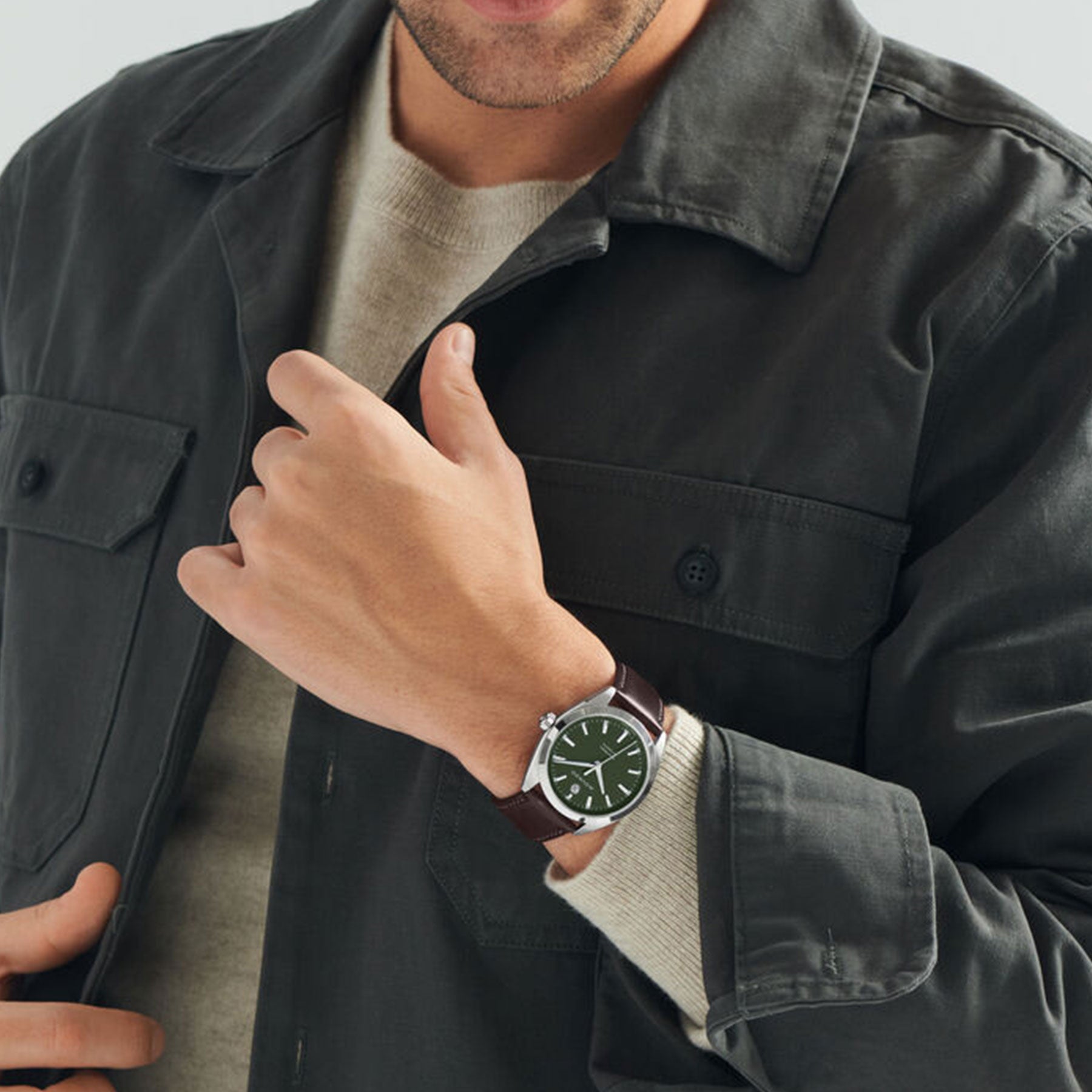 A person wearing a dark green jacket and beige sweater showcases the Movado Heritage Series Datron Automatic 40mm Watch on their wrist, featuring a green dial and brown leather strap. This Movado watch, equipped with an automatic movement, displays silver hour markers and hands. The neutral background highlights the elegance of its stainless steel case.