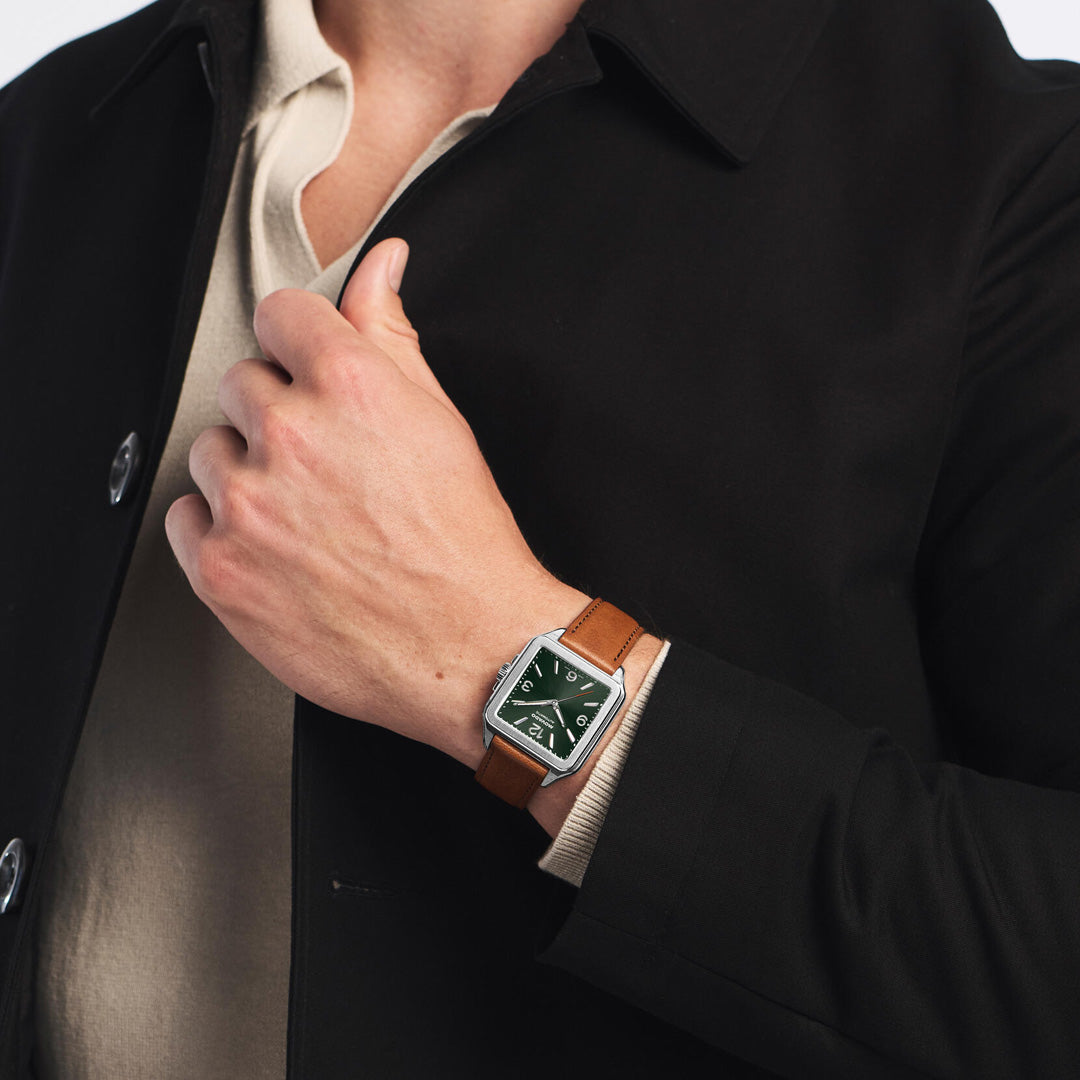 A person wearing a black coat and beige shirt shows their left hand, which features the Movado Heritage 1917 Automatic 44.6mm x 35mm Watch by Movado, an Art Deco timepiece with a square green face and brown leather strap.