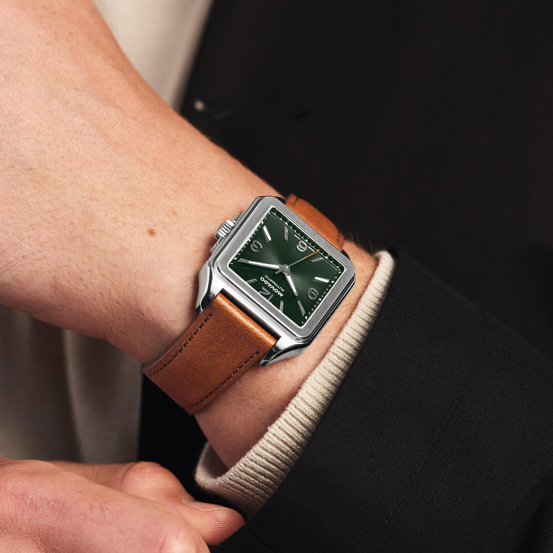 A close-up of a person wearing the Movado Heritage 1917 Automatic 44.6mm x 35mm watch by Movado—a square, green-faced wristwatch with a brown leather strap, styled with a black blazer over a beige sweater.