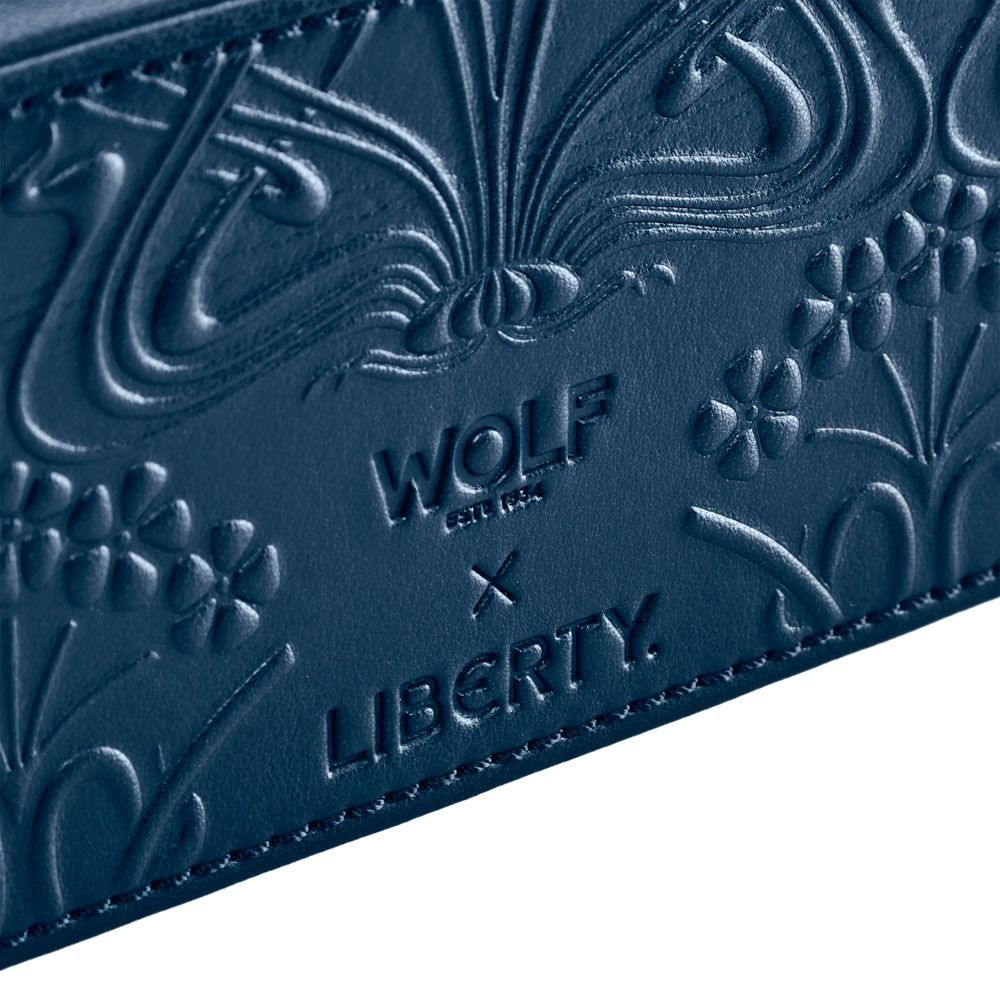 Close-up of embossed blue leather showcasing intricate floral and ornate Ianthe patterns, inspired by a luxurious WOLF 1834 x Liberty All Over Ianthe Medium Jewellery Zip Case in navy. The text reads: “WOLF EST. 1834 x LIBERTY.”.