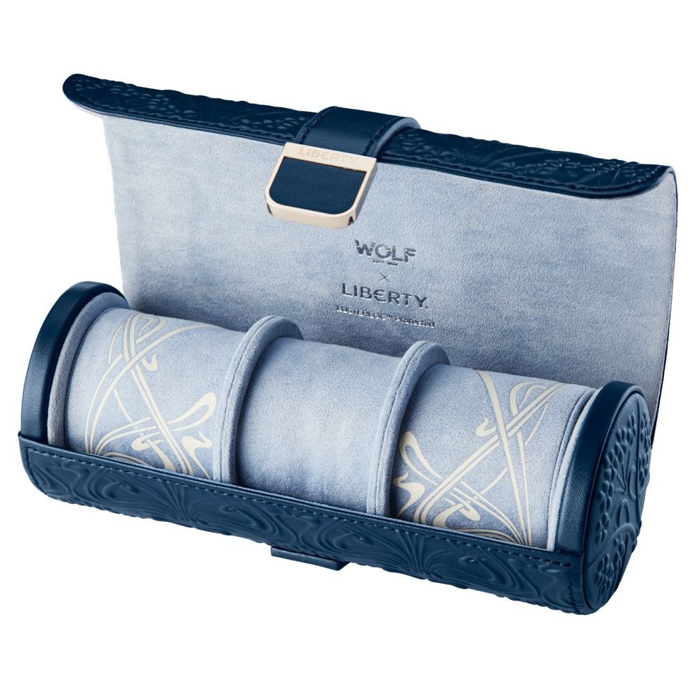 The WOLF 1834 X Liberty All Over Ianthe Triple Watch Roll Navy features intricate embossed designs in blue and light gray, a cylindrical shape reminiscent of a classic watch case, a gold buckle, and “WOLF Liberty” branding on the interior lining.