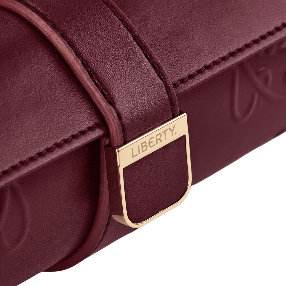 Close-up of the WOLF 1834 X Liberty All Over Ianthe Triple Watch Roll in Burgundy—a luxurious leather case by WOLF, featuring embossed patterns and a gold buckle engraved with “LIBERTY.” The elegant maroon leather evokes the sophistication of classic travel accessories, thoughtfully designed to protect your watches in style.