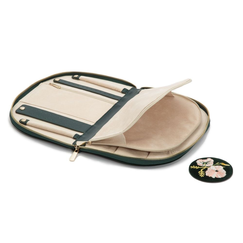 The WOLF 1834 Zoe Jewellery Portfolio, an open zippered beige and green case with compartments holding two pens, is shown next to a round patch embroidered with pink flowers and green leaves.