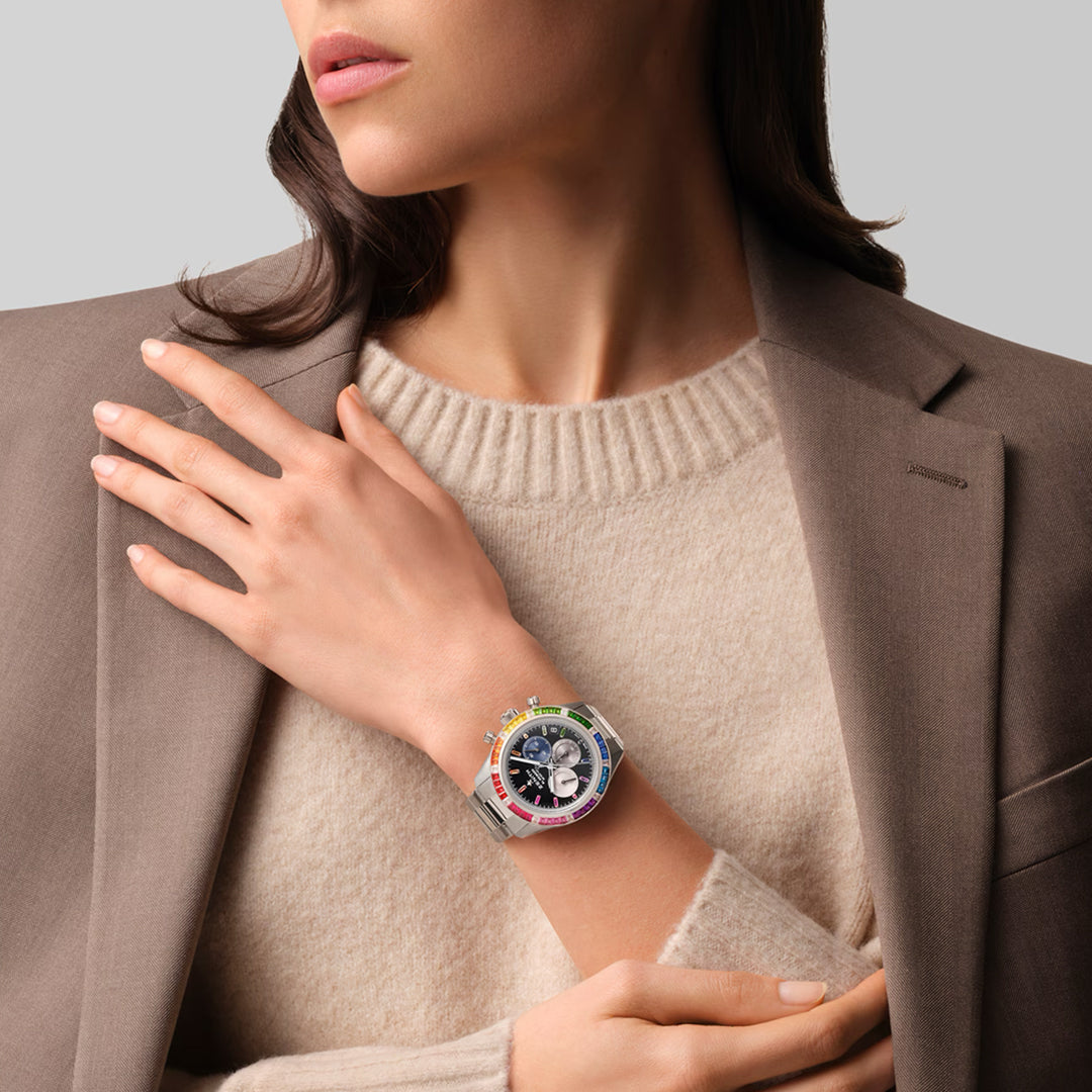 A woman in a light brown blazer and cream sweater stylishly displays the Zenith Chronomaster Sport El Primero 3600 41mm watch. The piece features rainbow sapphires in a white gold case, set against a plain gray background, with her hand gracefully on her shoulder.