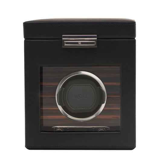 The WOLF 1834 Roadster Single Watch Winder With Storage is a chic black accessory featuring a transparent window that reveals its interior. It flaunts a vegan leather texture and metal accents, with a single circular slot designed for one watch. The brand name "WOLF" is prominently displayed inside, highlighting its patented design innovation.