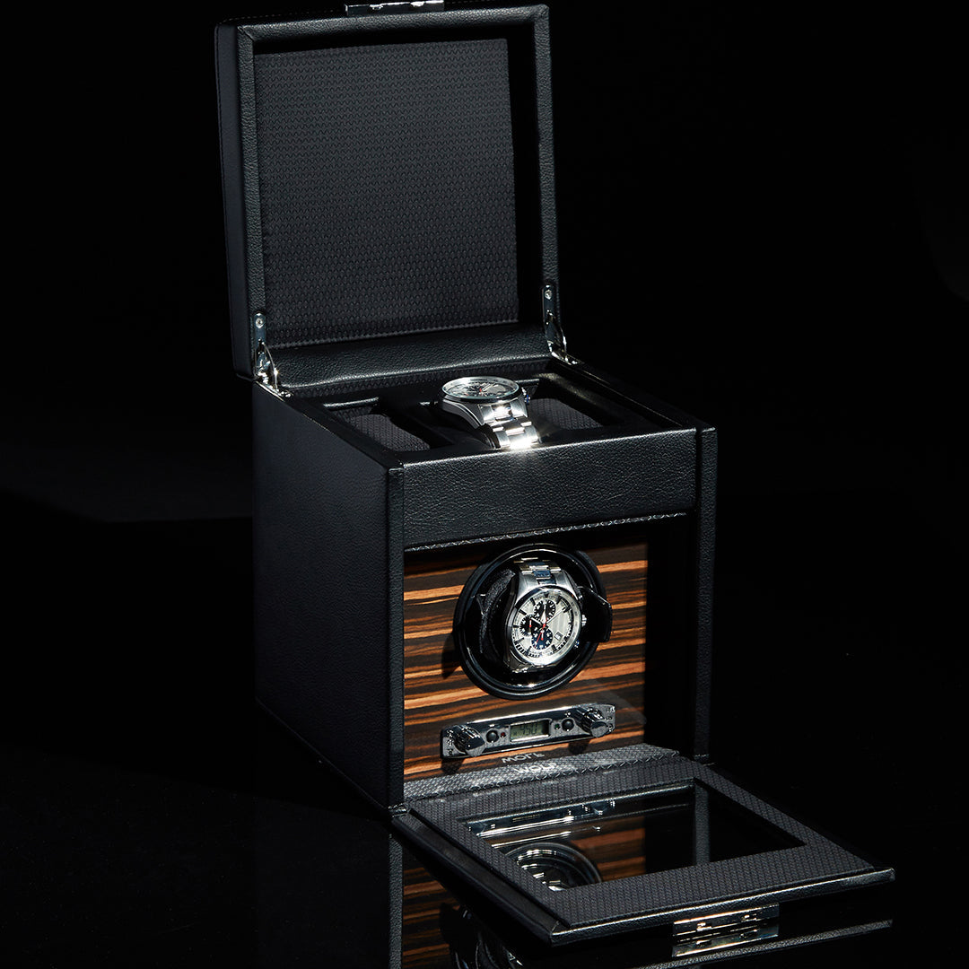 The WOLF 1834 Roadster Single Watch Winder With Storage is a luxury timepiece accessory from the brand WOLF. It boasts a sleek black and wood design with vegan leather accents. A silver watch is elegantly showcased through its open lid and transparent window, emphasizing the intricate, patented innovation within the winder's mechanism.