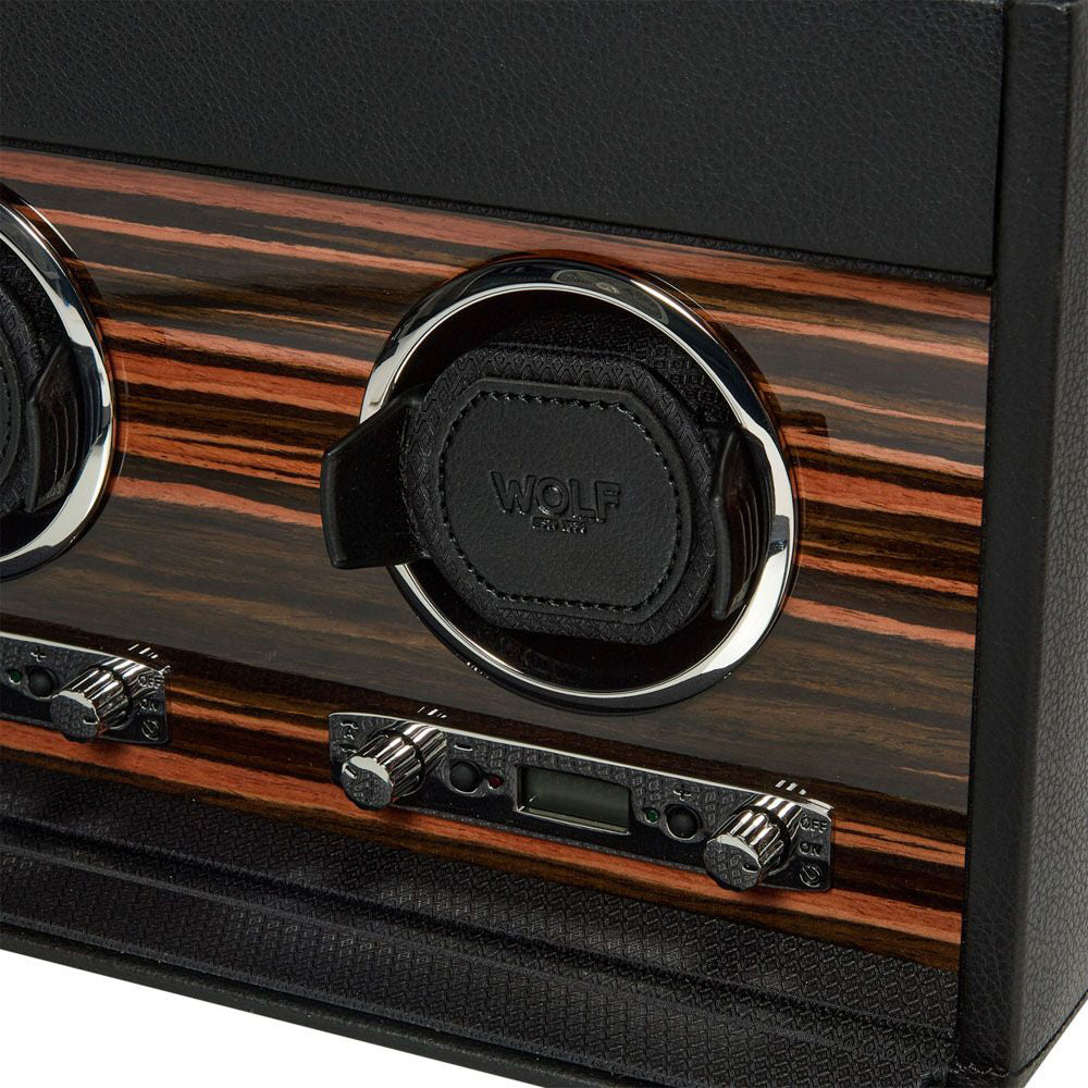 Close-up of the WOLF 1834 Roadster Triple Watch Winder With Storage, showcasing two circular compartments with gleaming rims against a striped wood panel. The compartments are labeled "WOLF" and feature black leather covers, each housing a dynamic cuff. Below, you'll find two adjustable dials and a small display for the power reserve setting.