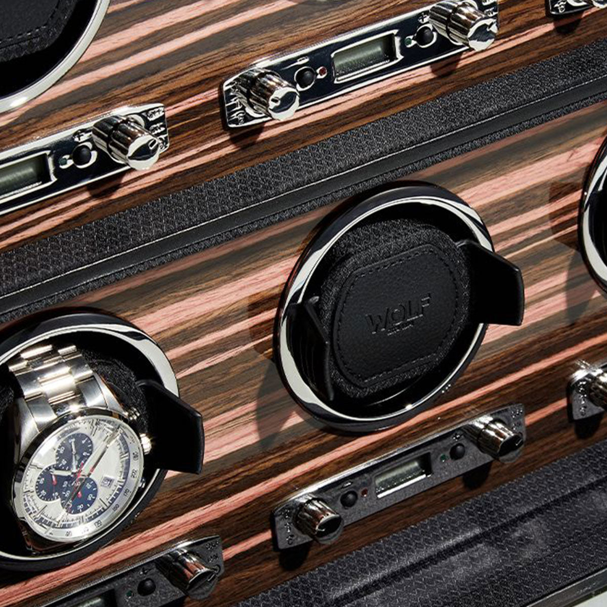 An intricate view of the WOLF 1834 Roadster 6 Piece Winder, featuring an ebony macassar wood finish with metallic accents, highlights its multiple sleek compartments. One compartment holds a stainless steel wristwatch, all complemented by smooth black holders.