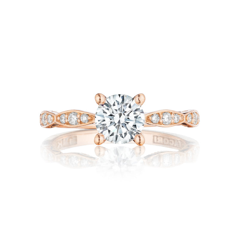 This Tacori Sculpted Crescent engagement ring, crafted in 18K rose gold, features a round brilliant solitaire at its center. The band is adorned with small diamonds arranged in intricate patterns on either side of the centerpiece, all beautifully reflecting on a white surface.