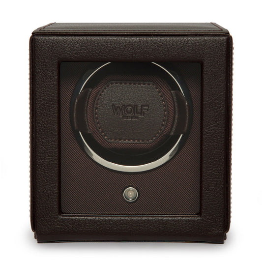 The WOLF 1834 Cub Single Watch Winder With Cover, crafted from brown vegan leather with a transparent front, prominently showcases the "Wolf" logo. Its interior features plush cushioning engineered to securely hold and rotate a watch using its patented rotation system. This sleek, cubic winder includes a conveniently positioned button at the bottom center for easy operation.