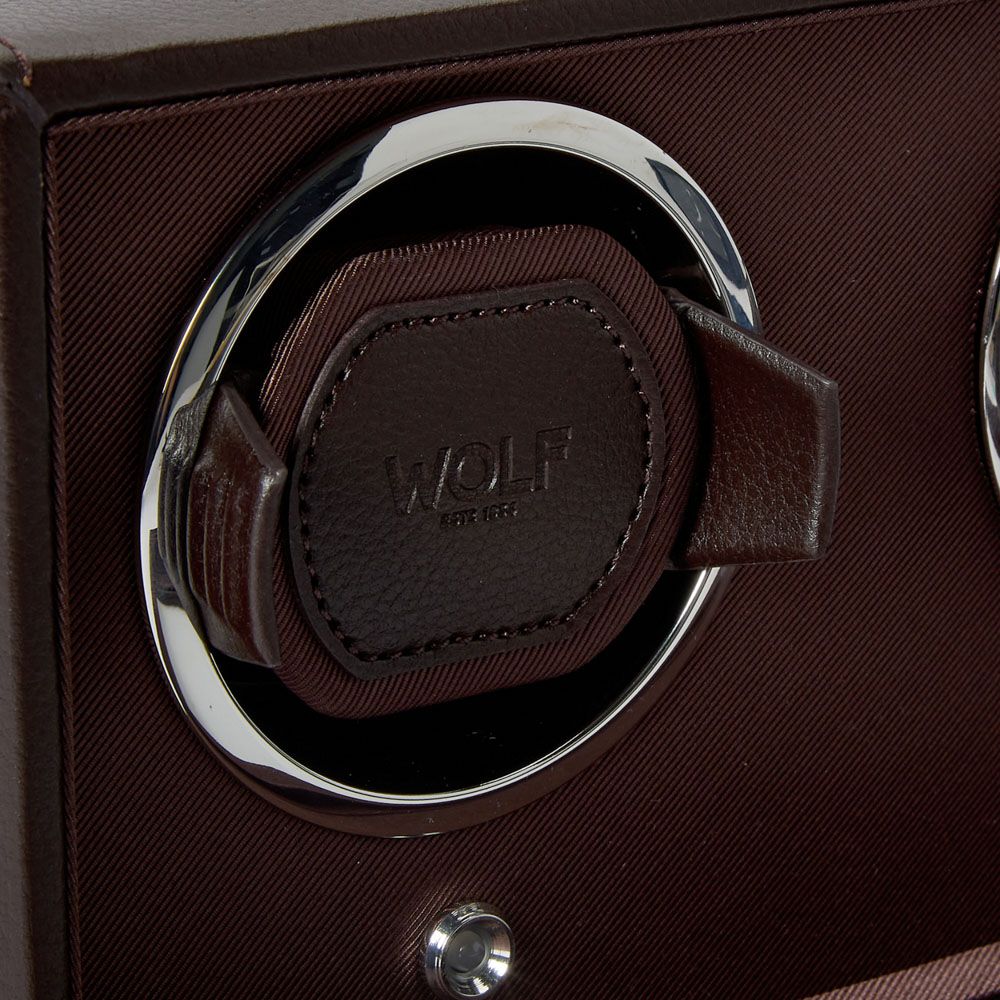 Close-up of the WOLF 1834 Cub Double Winder with Cover in Brown, crafted from brown vegan leather and set within a circular chrome frame, featuring the “WOLF EST. 1834” embossing. This elegant watch winder by WOLF provides advanced rotation programs to maintain your timepieces in perfect condition. The background includes textured brown fabric.