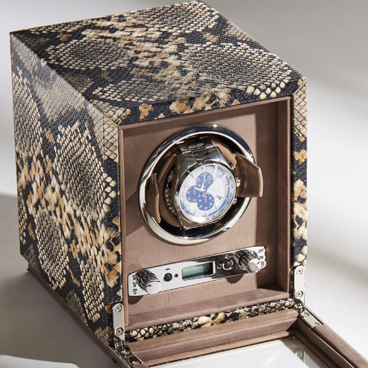 The WOLF 1834 Exotic Single Watch Winder, crafted by WOLF in top grain cowhide leather, elegantly showcases a silver watch with a blue and white face. Its open design reveals a digital interface and control knobs that ensure precise rotations.