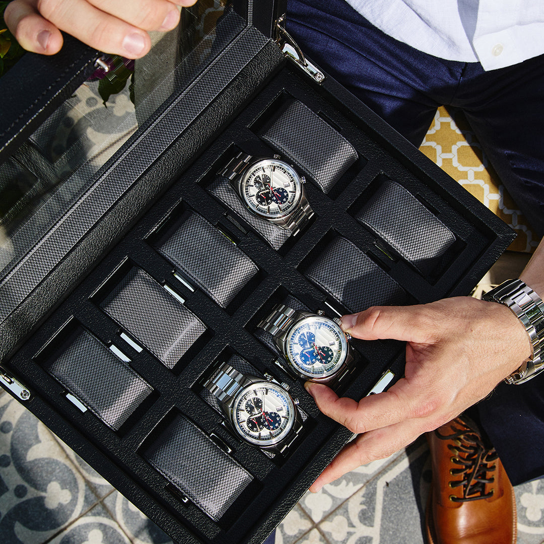 A person in a white shirt and brown leather shoes carefully adjusts a watch in the WOLF 1834 10 Piece Watch Box. This watch storage case by WOLF features a transparent lid and elegantly accommodates nine silver wristwatches with black dials, perfectly organized for those who appreciate both style and functionality.