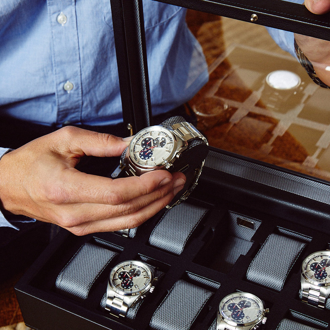 A person in a blue shirt is holding a luxury watch from the WOLF 1834 10 Piece Watch Box, crafted with vegan leather. The box, featuring a dark exterior and transparent lid, elegantly displays several other watches arranged neatly inside.