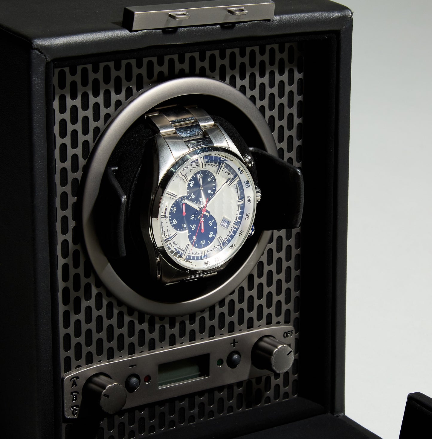 A wristwatch with a silver band and blue chronograph dial is elegantly displayed in the WOLF 1834 Axis Single Watch Winder. This winder from WOLF features control buttons, multiple rotation options, and a digital display located below the watch holder, along with a lock-in dynamic cuff for secure placement.