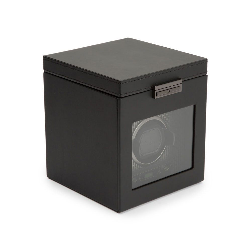 Introducing the WOLF 1834 Axis Single Watch Winder With Storage, a stylish square-shaped black box featuring a transparent front panel. Designed by WOLF, this exquisite watch winder offers various rotation options and includes a hinged lid with a metallic clasp on the top front edge. It also comes equipped with a lock-in dynamic cuff to ensure secure storage for your timepiece.