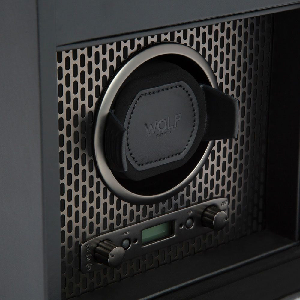Close-up of a black WOLF 1834 Axis Single Watch Winder With Storage set against a grid pattern background. This winder accommodates one watch and includes a digital display, control knobs, and a lock-in dynamic cuff for added security. The brand "WOLF" is embossed on the holder, providing versatile rotation options.