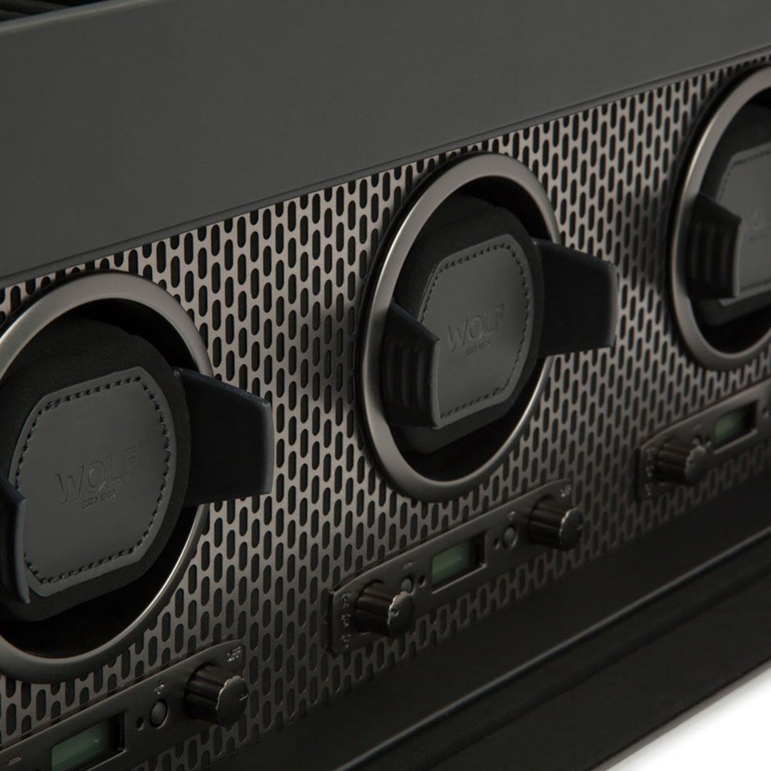 Close-up of the WOLF 1834 Axis Triple Watch Winder With Storage, a modern device with a sleek design showcasing three circular components with textured grips set on a perforated metallic panel. Below each component are knobs and digital displays that evoke the precision of a watch winder.